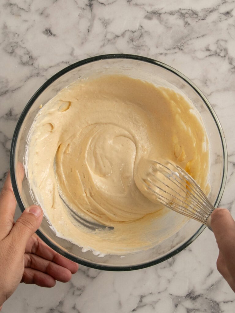 A person holding a glass bowl with creamy batter inside, whisking it with a metal whisk on a marble countertop.