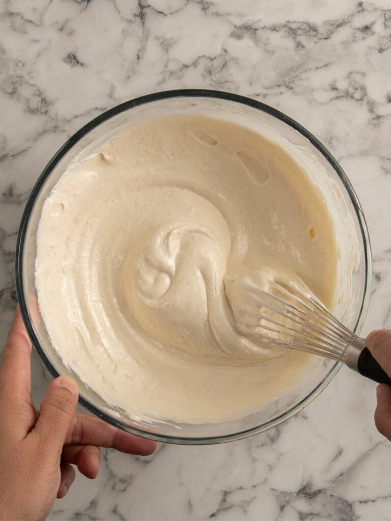 A hand holds a glass bowl filled with creamy batter while another hand whisks it on a white marble countertop.