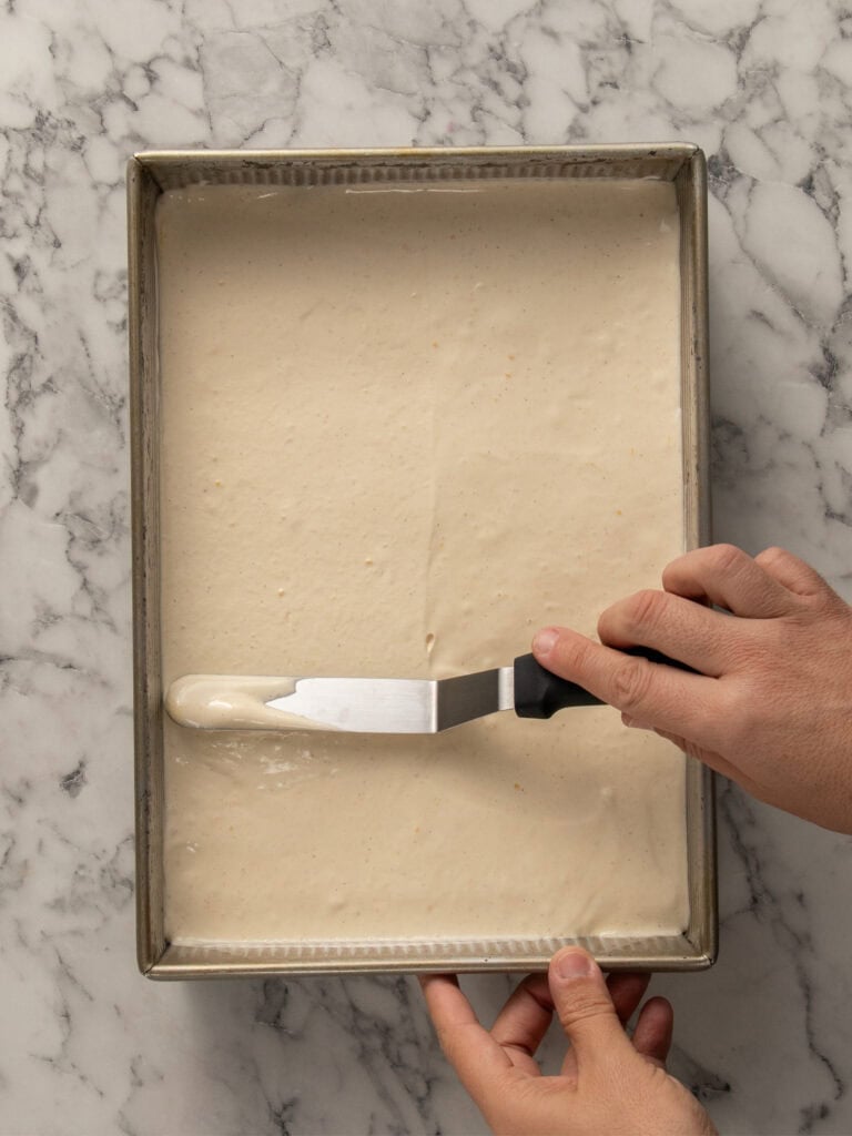 A person uses an offset spatula to spread cake batter evenly in a rectangular metal baking pan on a marble surface.