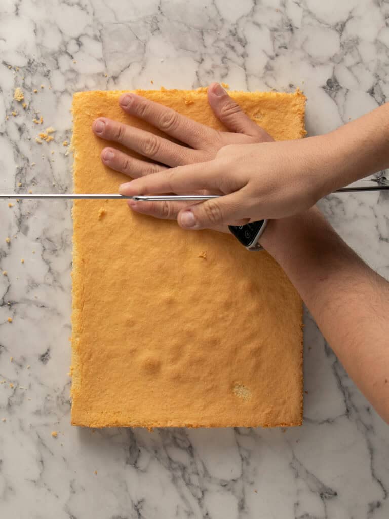 A hand holds a rectangular sponge cake steady on a marble surface while another hand slices it horizontally with a long serrated knife.