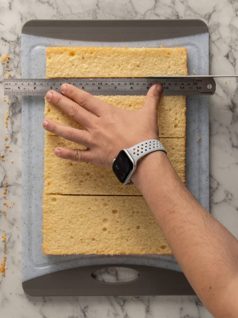 A hand holds a ruler to measure a rectangular sponge cake on a cutting board; the cake is being cut into sections on a marble countertop.