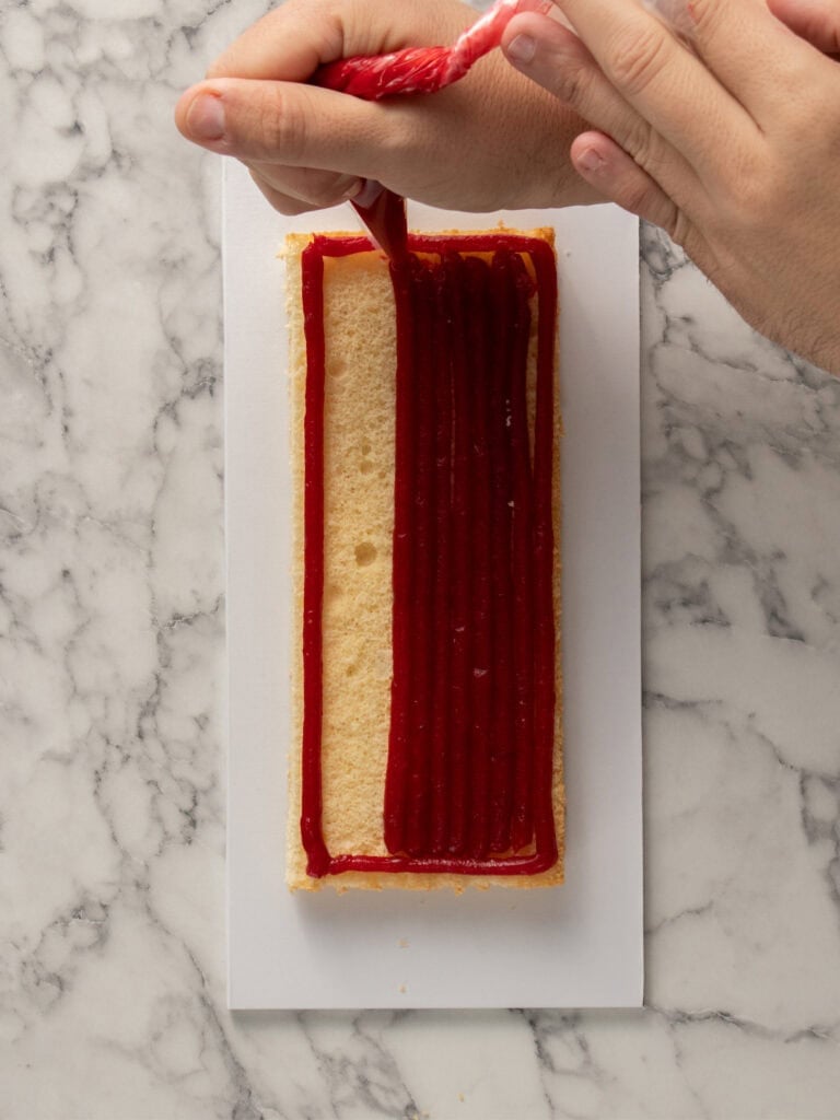 A hand is piping red jam in lines onto a rectangular layer of cake on a white surface with a marble background.