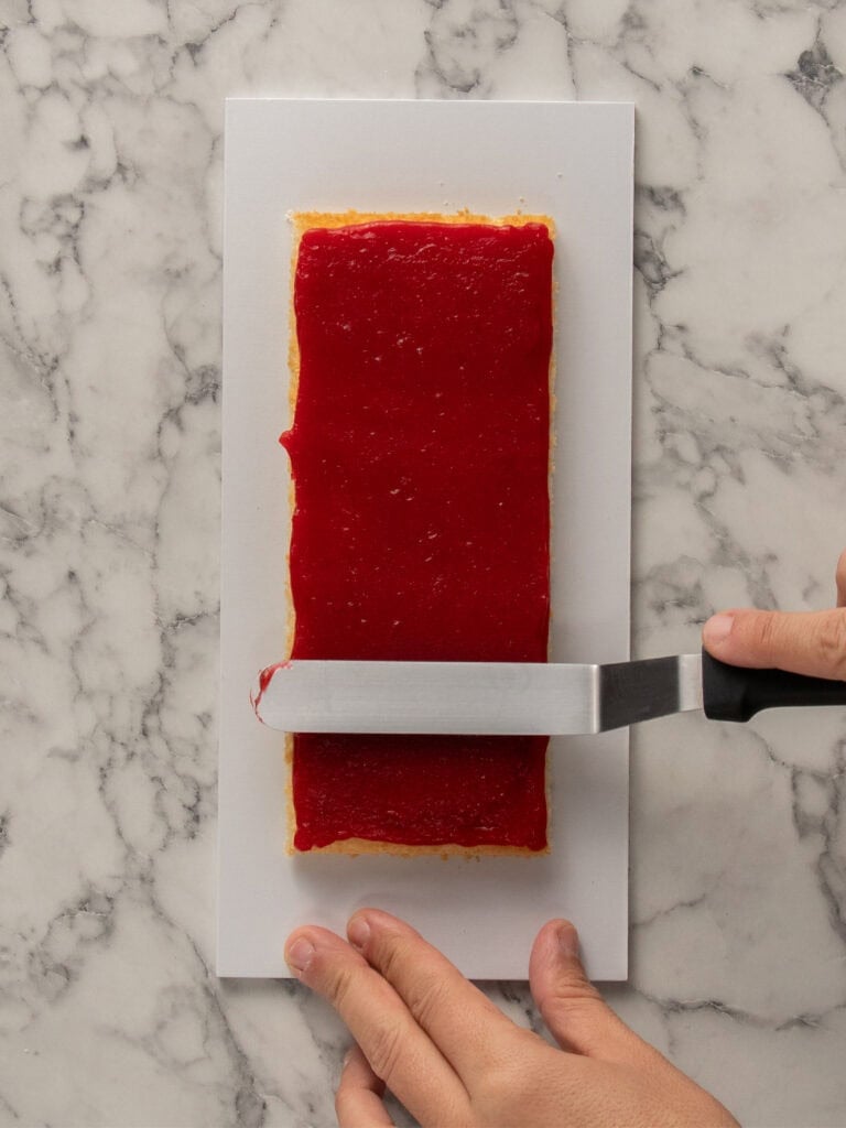A hand uses an offset spatula to spread a smooth, bright red layer of fruit jam over a rectangular pastry on a white rectangular plate, placed on a marble countertop.