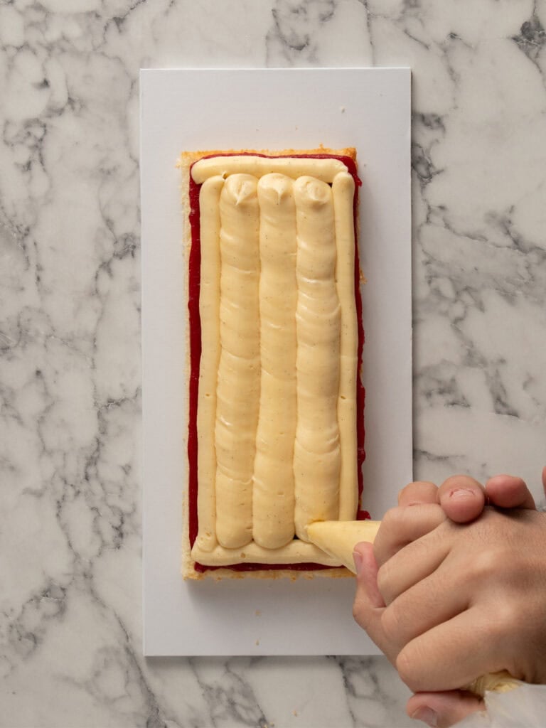 A hand uses a piping bag to add thick lines of pastry cream onto a rectangular pastry base with a red jam layer, all on a white rectangular board atop a marble surface.