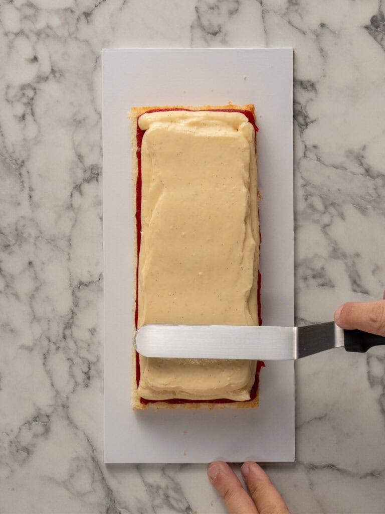 A hand uses an offset spatula to spread pastry cream over a rectangular cake layer with a red jam filling, on a white cutting board set on a marbled surface.