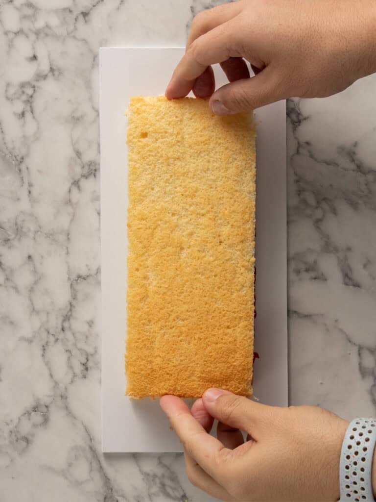 Two hands are placing a rectangular sponge cake layer onto a white rectangular board on a marble countertop.