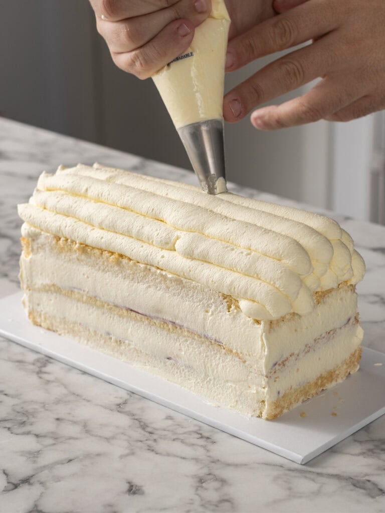 A person decorates a rectangular layered cake with piped Chantilly cream using a pastry bag on a marble countertop.