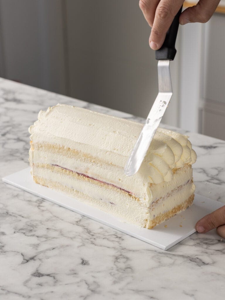 A person holds a large spatula and smooths Chantilly cream on a rectangular layer cake with cream and jam filling, set on a marble countertop.