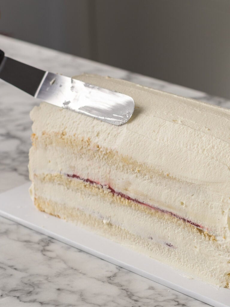 A cake with layers of pastry cream and jam is being frosted with Chantilly cream using an offset spatula on a marble countertop.
