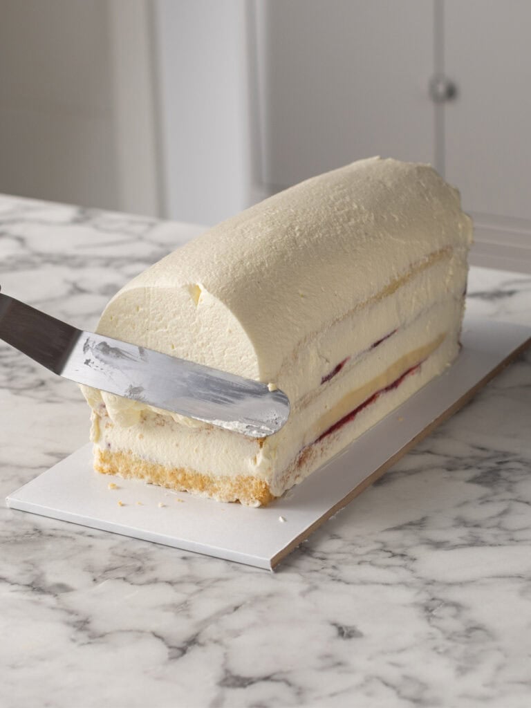 A rectangular layered cake with Chantilly cream is being smoothed on top with a metal spatula on a marble countertop.