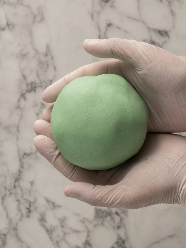 Gloved hands holding a smooth, round ball of light green marzipan against a white marble background.