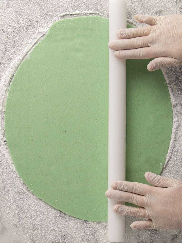 Gloved hands use a white rolling pin to flatten a round sheet of green marzipan on a floured marble surface.