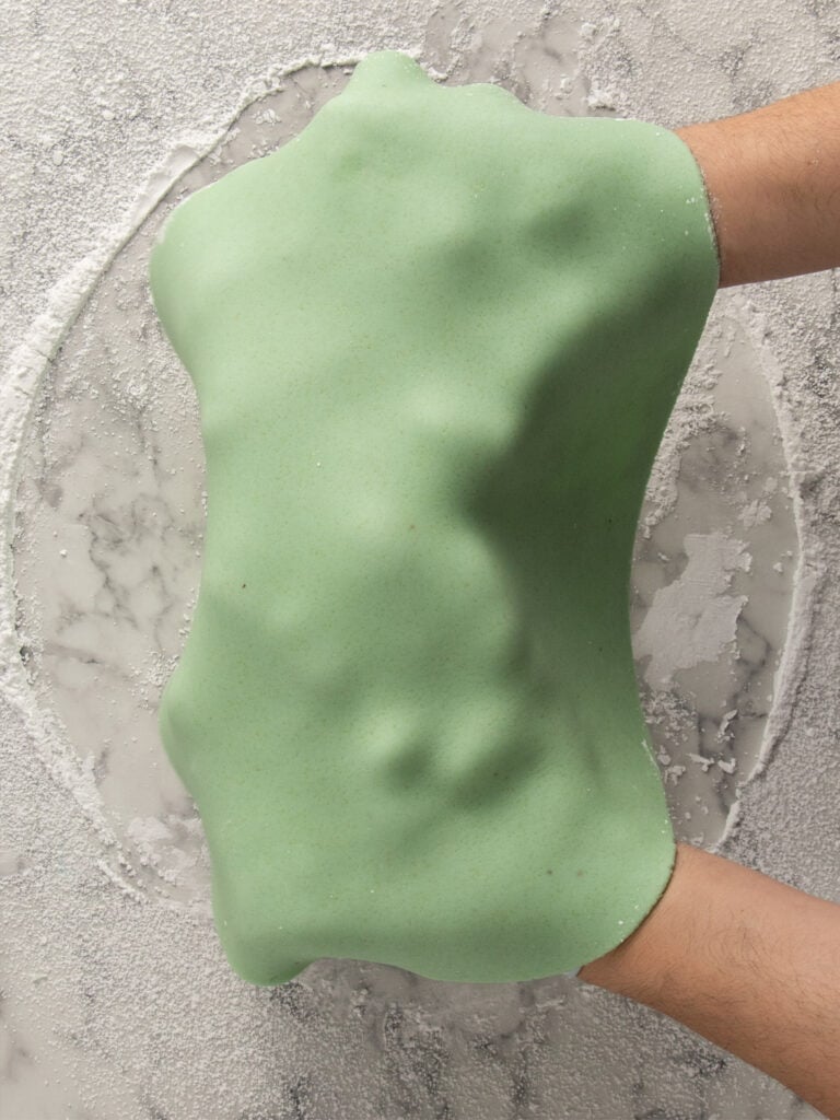 Two hands stretch a sheet of light green marzipan on a floured marble surface, showing its elasticity and smooth texture.