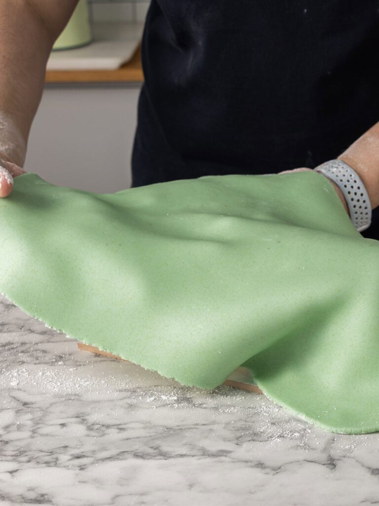 A person drapes a sheet of green marzipan over a cake on a marble countertop, preparing to cover it. Flour is scattered on the counter.