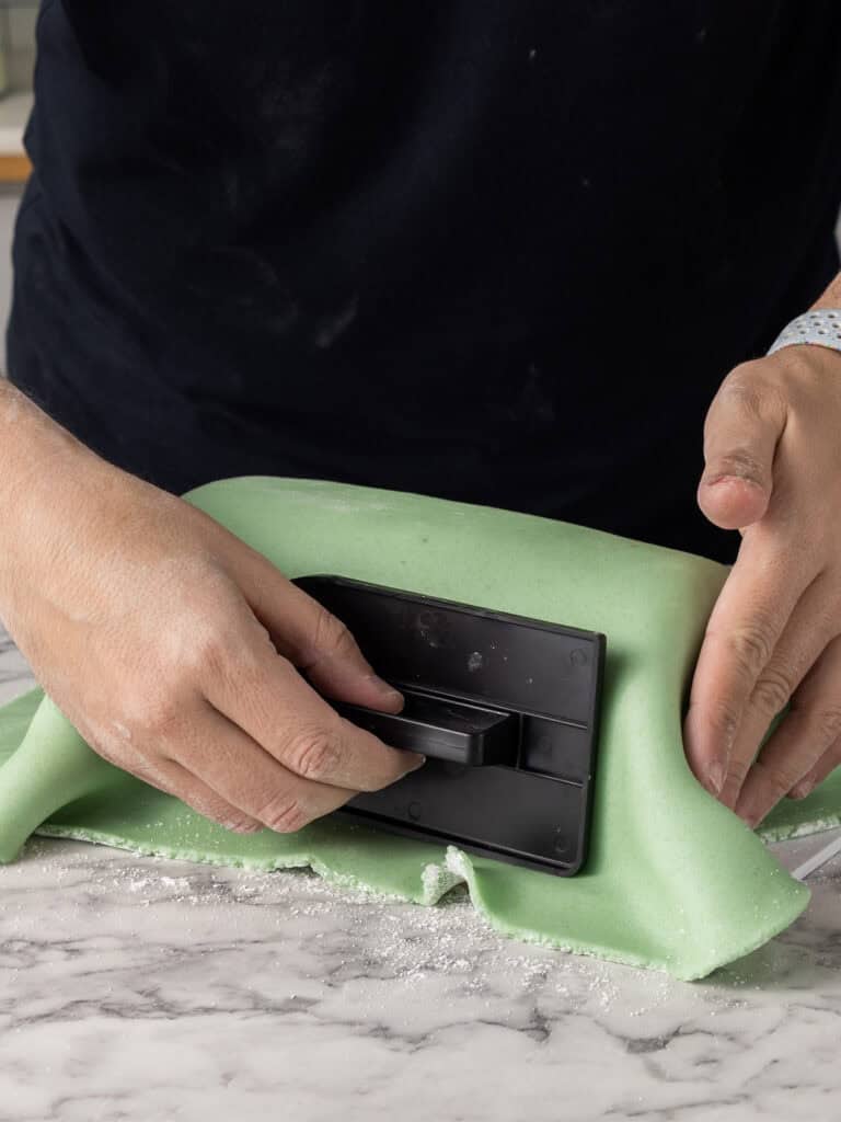 A person smooths green marzipan over a cake on a marble countertop using a black fondant smoother, with powdered sugar scattered on the surface.