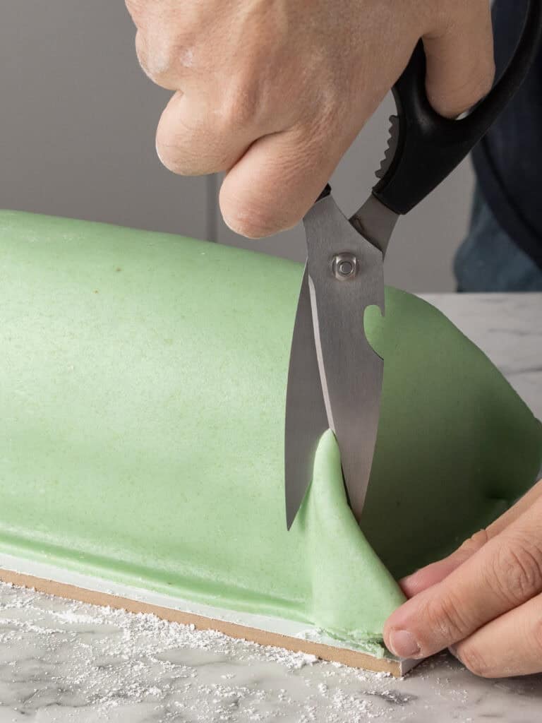A person uses kitchen scissors to trim a sheet of pale green marzipan covering a cake on a marble surface. One hand holds the scissors, while the other holds the fondant in place.