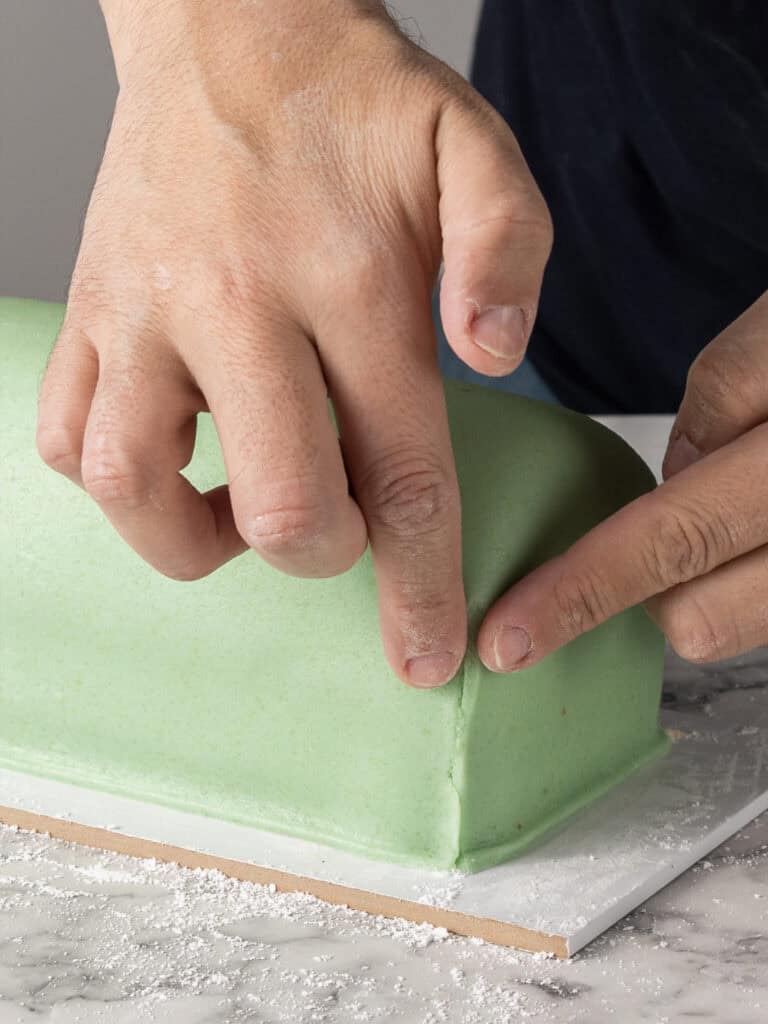 A person’s hands carefully smooth the edge of a rectangular cake covered in light green marzipan, on a white board dusted with powdered sugar.