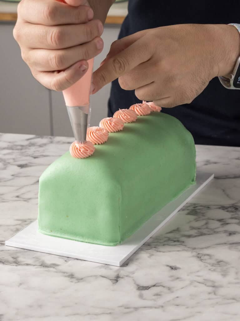 A person decorates a rectangular, green marzipan-covered cake with pink frosting swirls using a piping bag on a marble countertop.