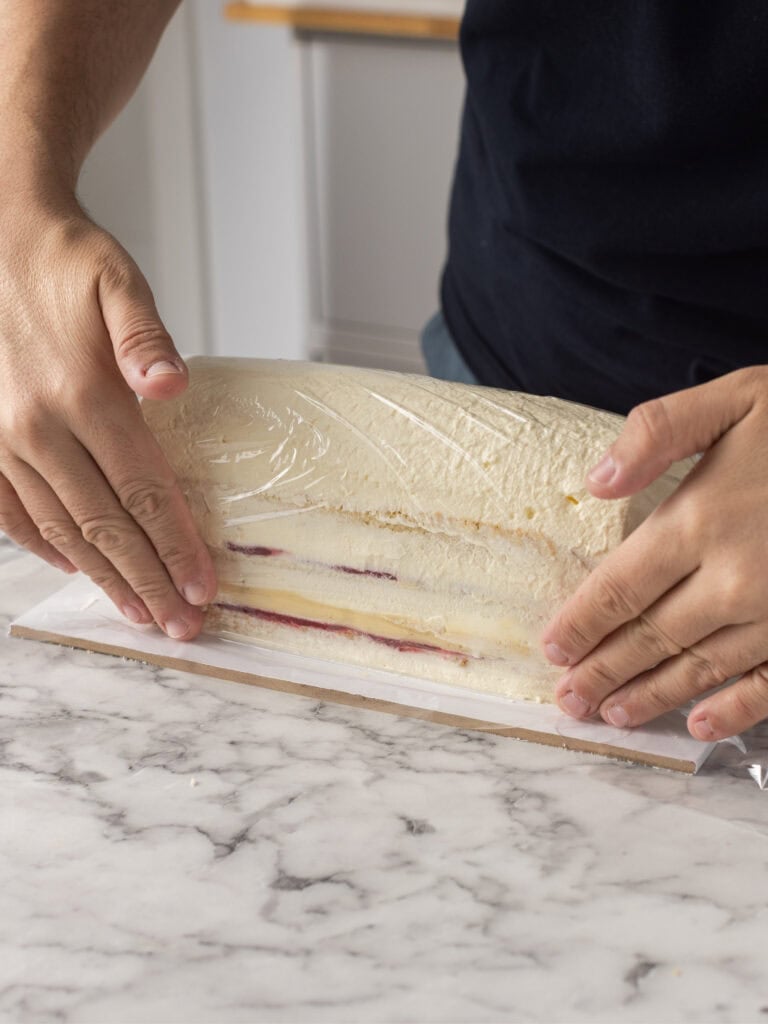 A person carefully lifts a layered dessert wrapped in plastic wrap onto a parchment-lined surface. The dessert has visible layers of cream and a red filling, set on a marble countertop.