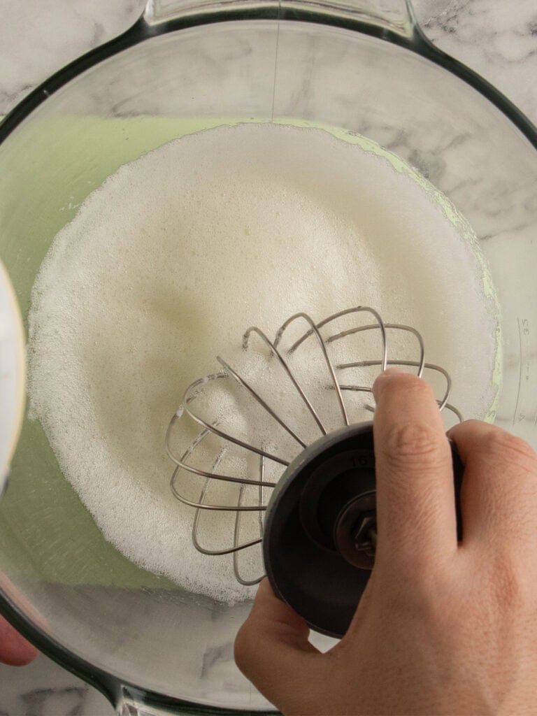 A hand holding a whisk attachment over a mixing bowl with frothy egg whites inside, viewed from above on a marble countertop.