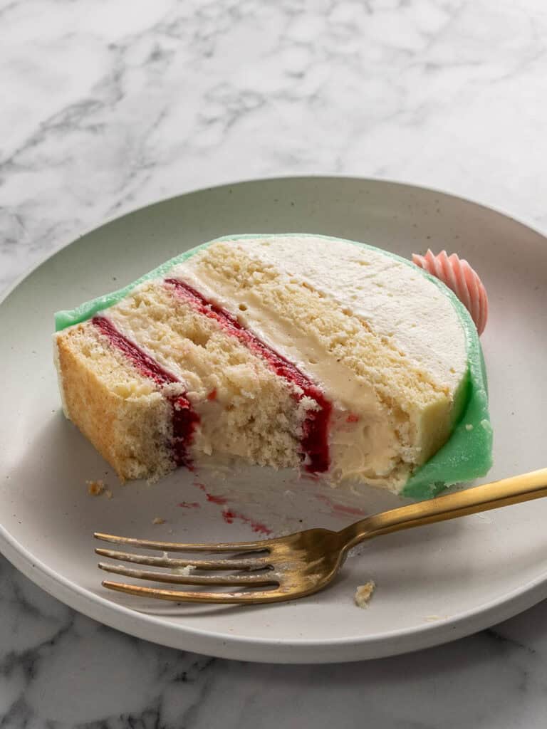 A slice of Swedish princess cake with white frosting and a red fruit filling sits on a white plate. A gold fork rests in front of the cake, and crumbs are scattered on the plate atop a marble surface.