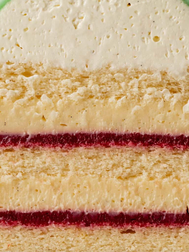 Close-up of a Swedish princess cake slice showing vanilla sponge cake, creamy frosting, and two thin layers of red fruit filling.