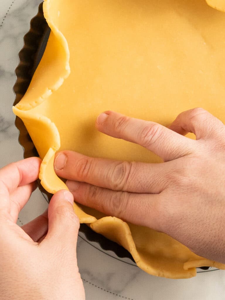 Hands gently pressing and shaping raw pie dough into a round, fluted tart pan on a marble countertop.
