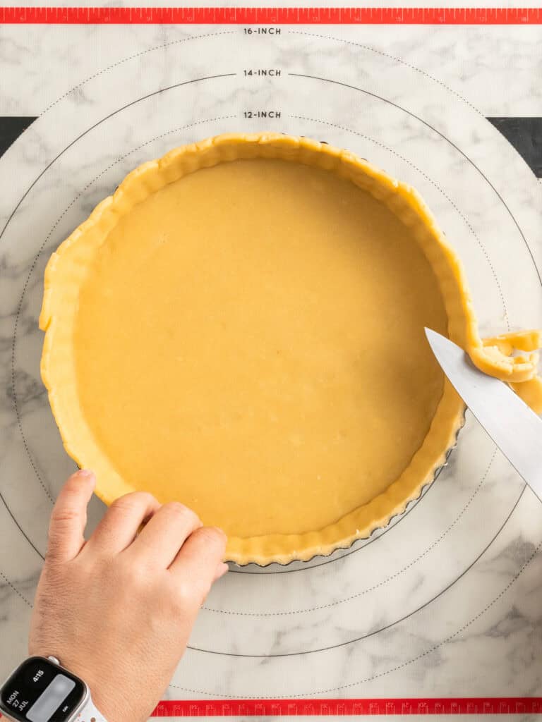A hand trims excess pastry dough from a tart pan using a knife. The tart pan sits on a marble surface with a red-bordered pastry mat marked with measurements.