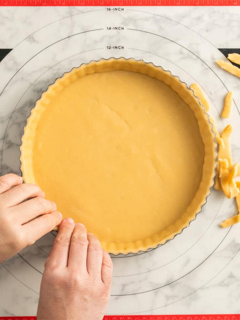 Hands pressing tart dough into a round tart pan on a marble surface with a pastry mat underneath; excess dough is being trimmed from the edges.