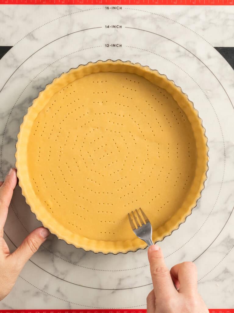 A person uses a fork to poke holes in an unbaked tart crust placed in a fluted tart pan on a marble surface marked with measurement guides.