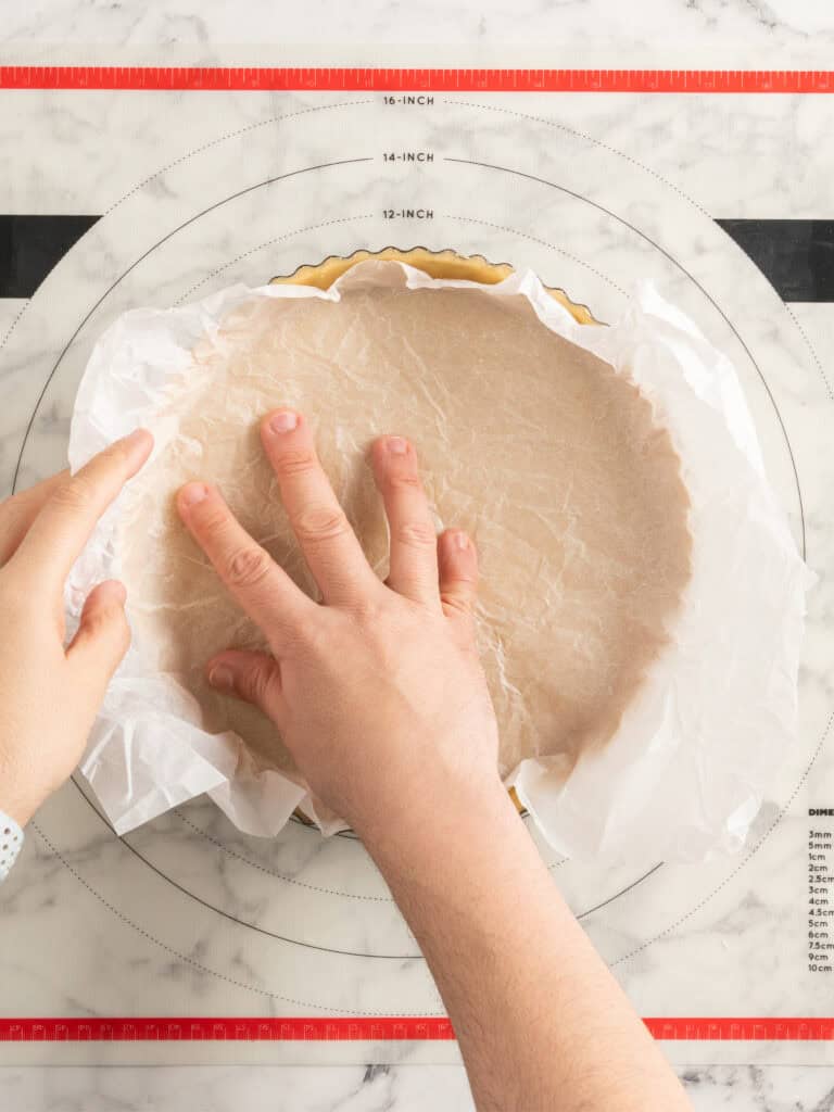 Hands pressing parchment paper into a pie crust on a round baking mat with measurement markings, on a marble counter.