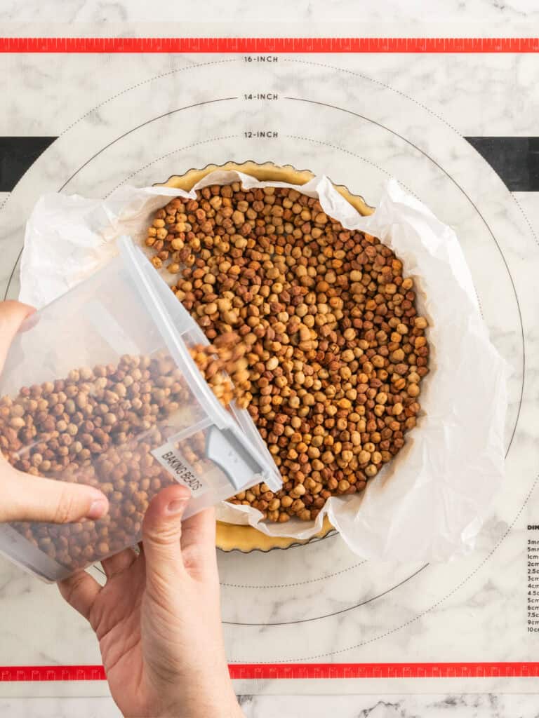 A person pours dried chickpeas from a container onto parchment paper in a pie crust for blind baking, with a pastry mat underneath.