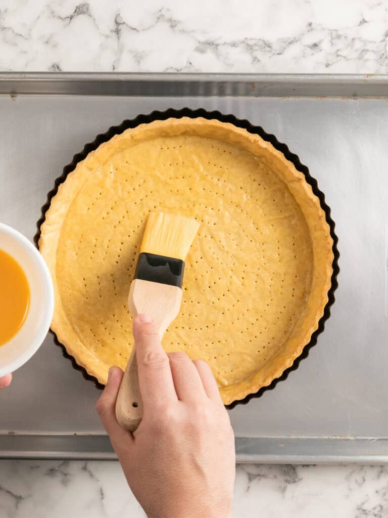 A hand brushes egg wash onto a raw, fork-pricked pie crust in a tart pan, which sits on a metal baking sheet on a marble countertop.