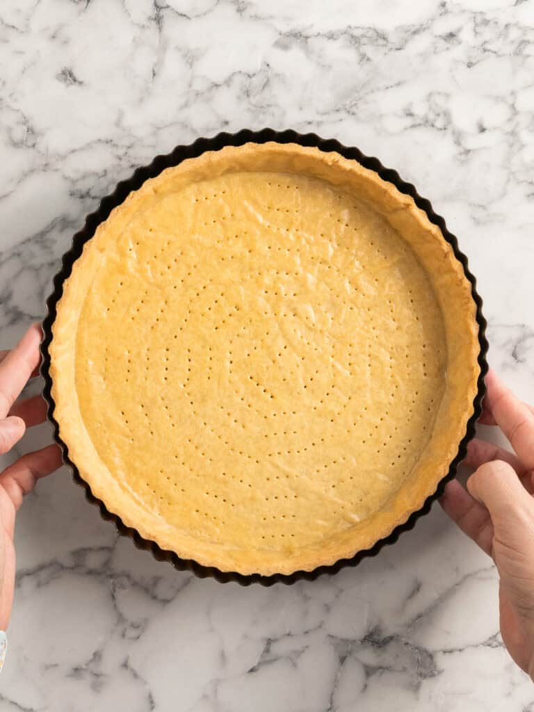 A person holds a round tart pan with an unbaked pie crust that has been pricked with a fork, on a marble countertop.