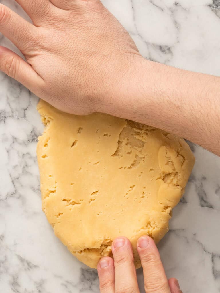 Two hands knead a piece of cookie dough on a white marble surface.