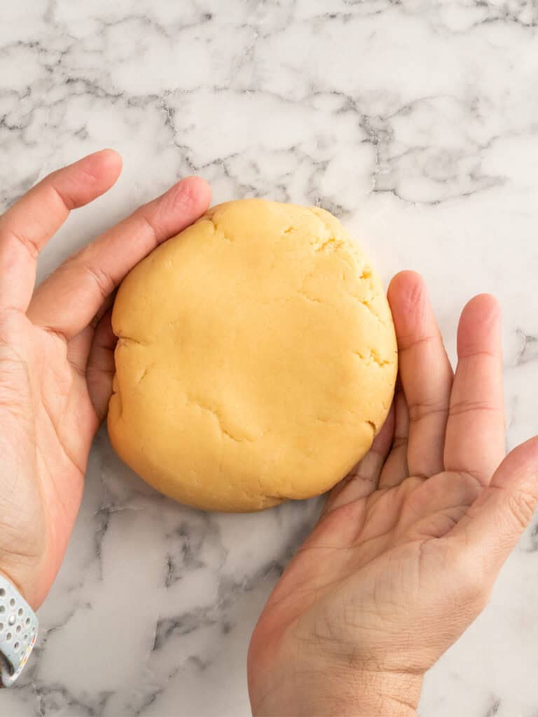 Two hands hold a round, smooth disc of dough on a white and gray marble countertop.