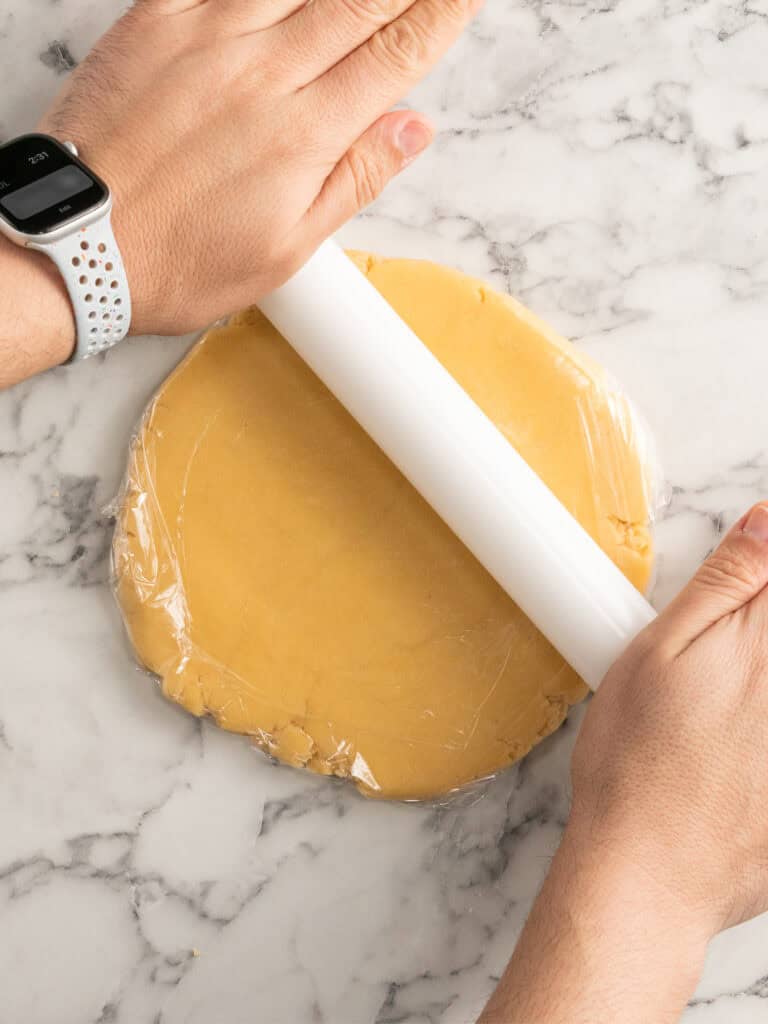 Hands rolling out cookie dough wrapped in plastic wrap on a marble surface with a white rolling pin. One wrist is wearing a white smartwatch.