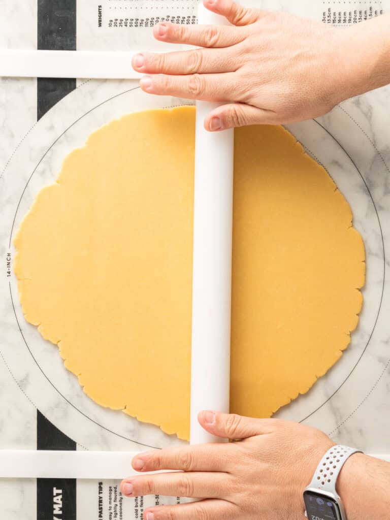 Hands rolling out dough with a rolling pin on a floured surface, using guides for even thickness. A smartwatch is visible on one wrist.