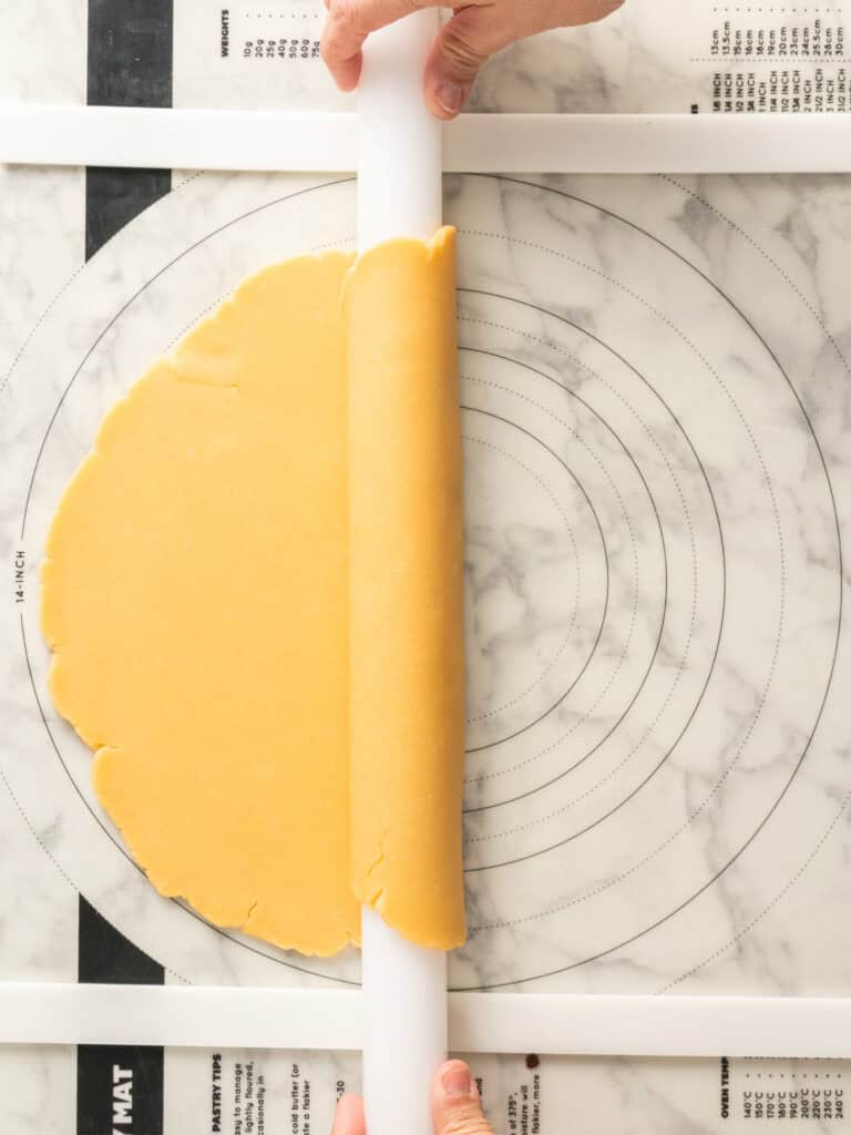 Hands use a rolling pin to roll out dough on a floured surface with circular measurement guides. The dough is partially flattened and spread in a rough oval shape.