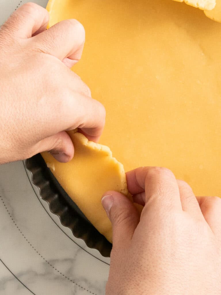 Hands pressing pie dough into a round tart pan on a light surface, shaping the edge of the crust before baking.
