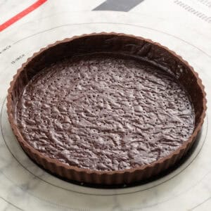 Unbaked chocolate tart crust in a fluted tart pan, shown on a circular baking mat with measurements, ready for filling or baking.