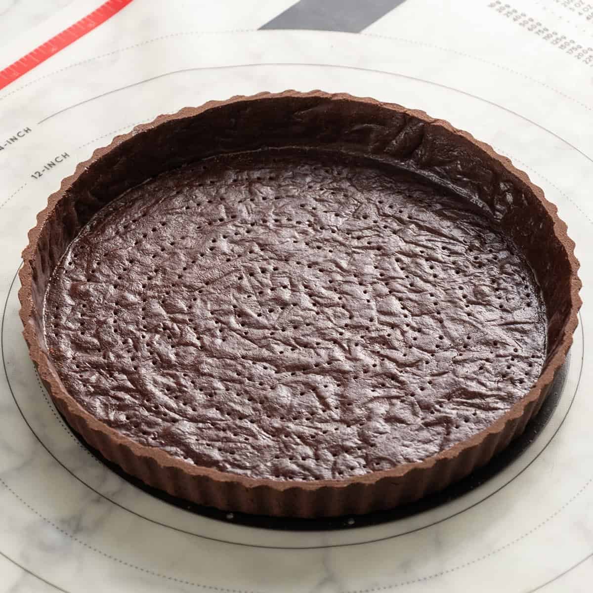 Unbaked chocolate tart crust in a fluted tart pan, shown on a circular baking mat with measurements, ready for filling or baking.