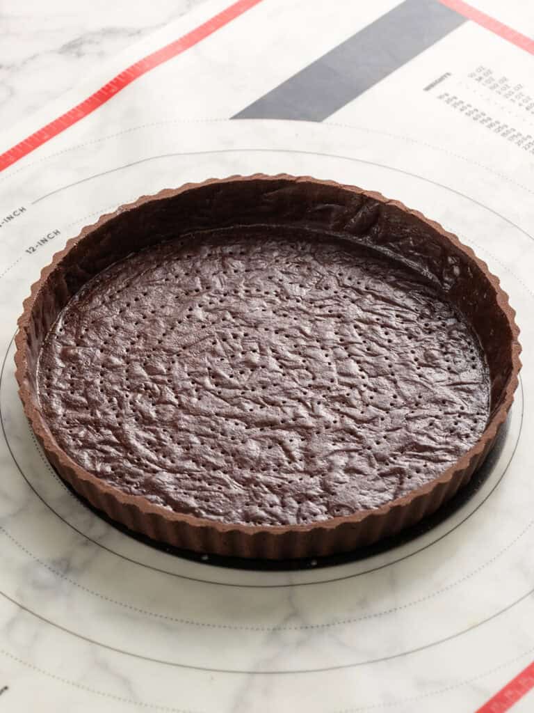 A chocolate tart crust with fork marks is in a fluted tart pan, placed on a marble surface with a baking mat underneath.