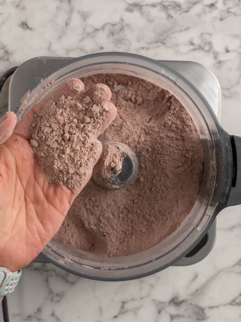 A hand holds a scoop of light brown powder above a food processor filled with the same powder, on a marble countertop.