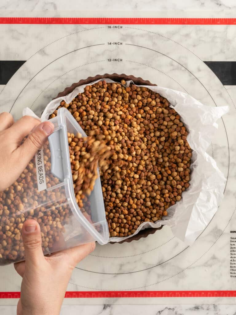 A person pours baking beads onto a parchment-lined tart crust in a pan placed on a marble surface with a circular measurement guide.