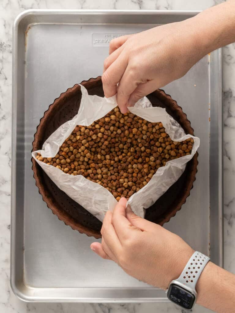 Hands lifting parchment paper filled with baking beans from a tart crust on a baking tray, preparing the crust for baking.