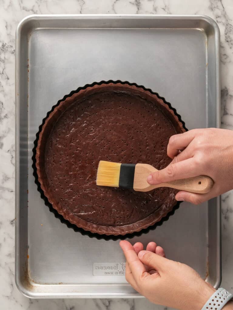 A person brushes a chocolate tart crust with a pastry brush, holding the tart pan over a metal baking sheet on a marble countertop.