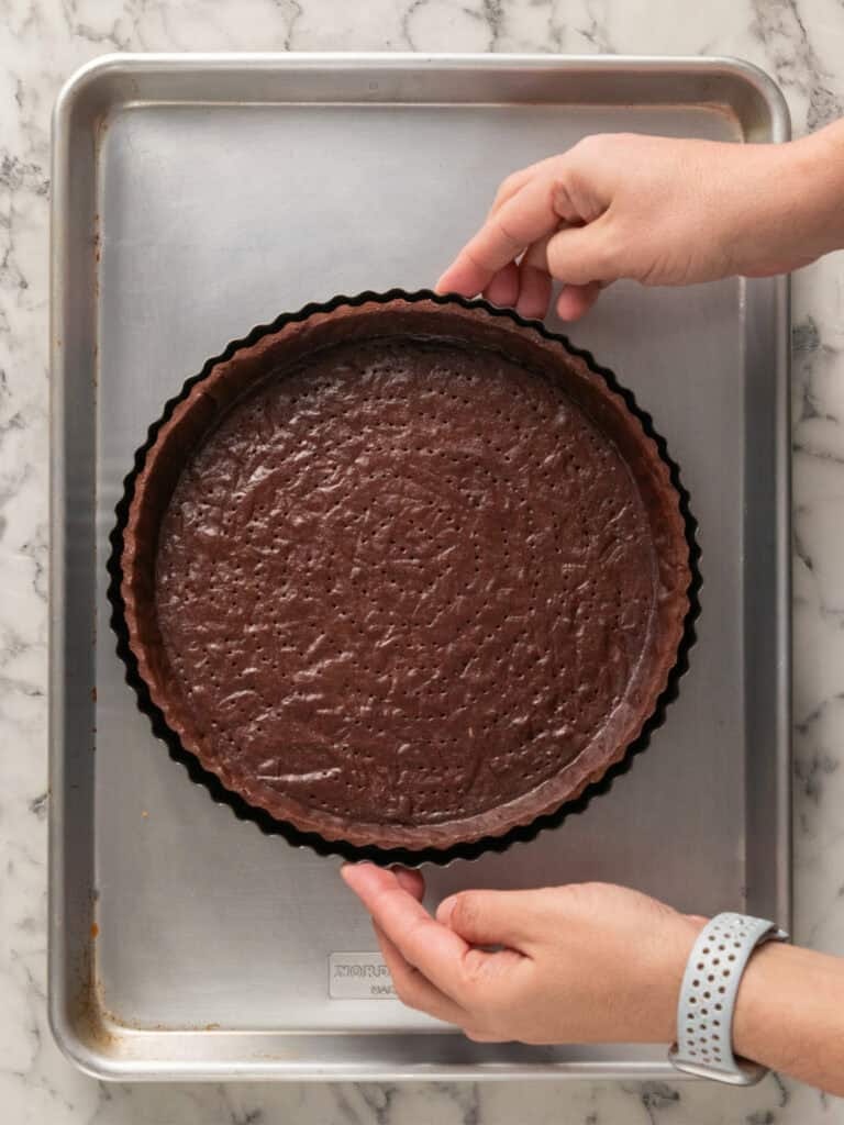 Hands holding a round tart pan filled with chocolate batter on a silver baking sheet atop a marble countertop.