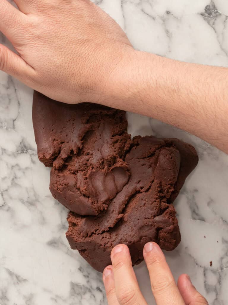 Hands kneading chocolate dough on a white marble surface.