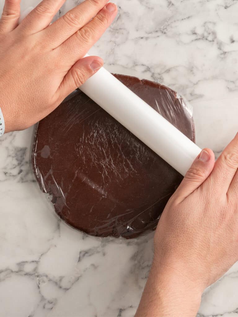 Two hands use a rolling pin to flatten chocolate dough covered in plastic wrap on a white marble surface.
